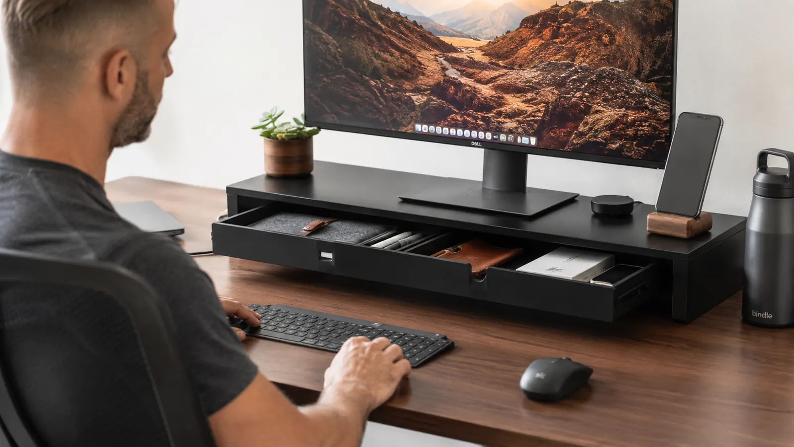 Clean home office desk with monitor elevated on a wooden-look riser, drawer slightly open revealing small accessories inside, warm ambient lighting
